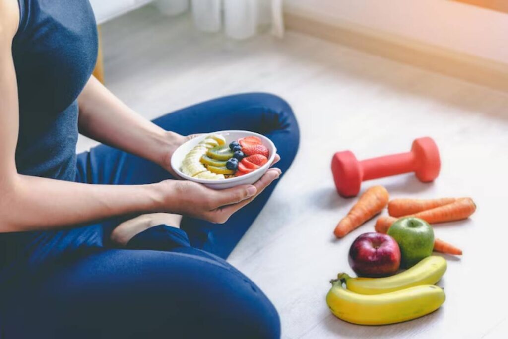 A person sits cross-legged, holding a bowl of fruit, surrounded by fresh vegetables and a red dumbbell, promoting healthy living.