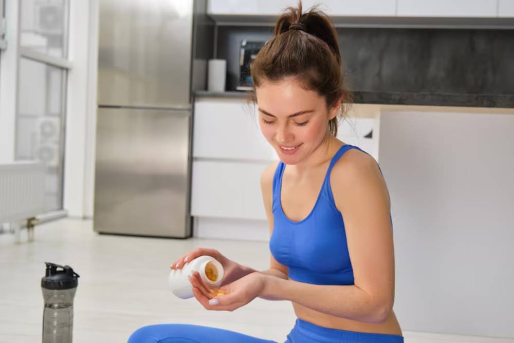 A woman in a blue athletic outfit sitting on the floor, holding a container of supplements, with a water bottle nearby in a modern kitchen.