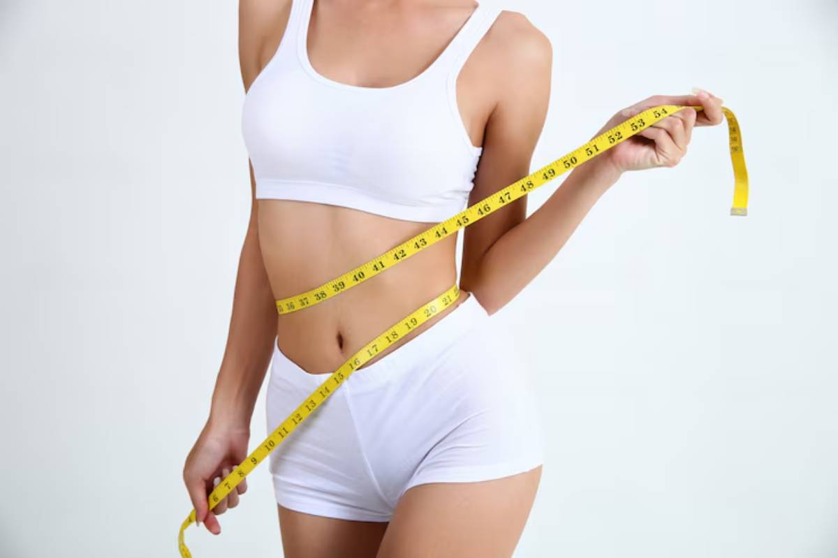 A woman in white sportswear holds a yellow measuring tape around her waist, suggesting fitness and a focus on body measurements. 