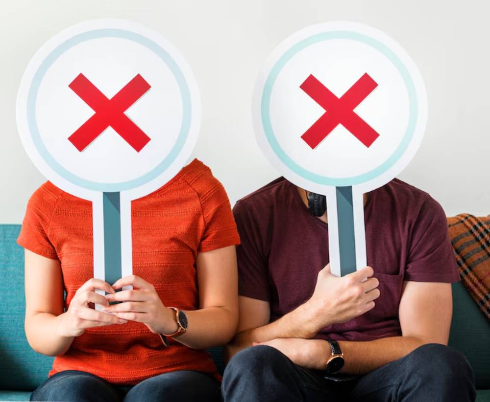 A woman and man sitting together on a couch hold large signs with red crosses, covering their faces, indicating disapproval.