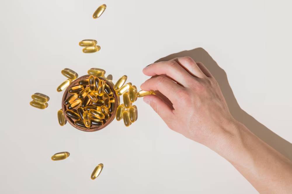 A hand reaching for a golden capsule above a bowl filled with more capsules, scattered on a light background.