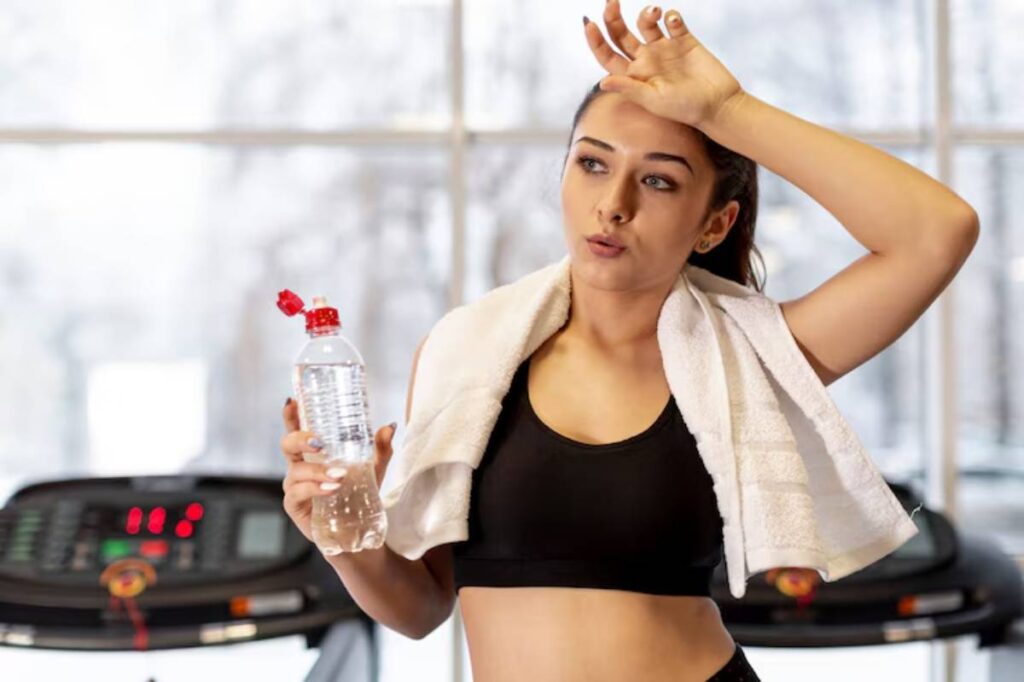A fit individual in a black sports bra, holding a water bottle, with a towel draped around their neck, stands in a gym setting.
