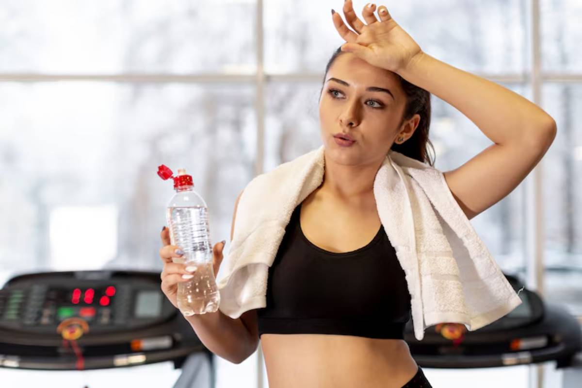 A fit individual in a black sports bra, holding a water bottle, with a towel draped around their neck, stands in a gym setting.