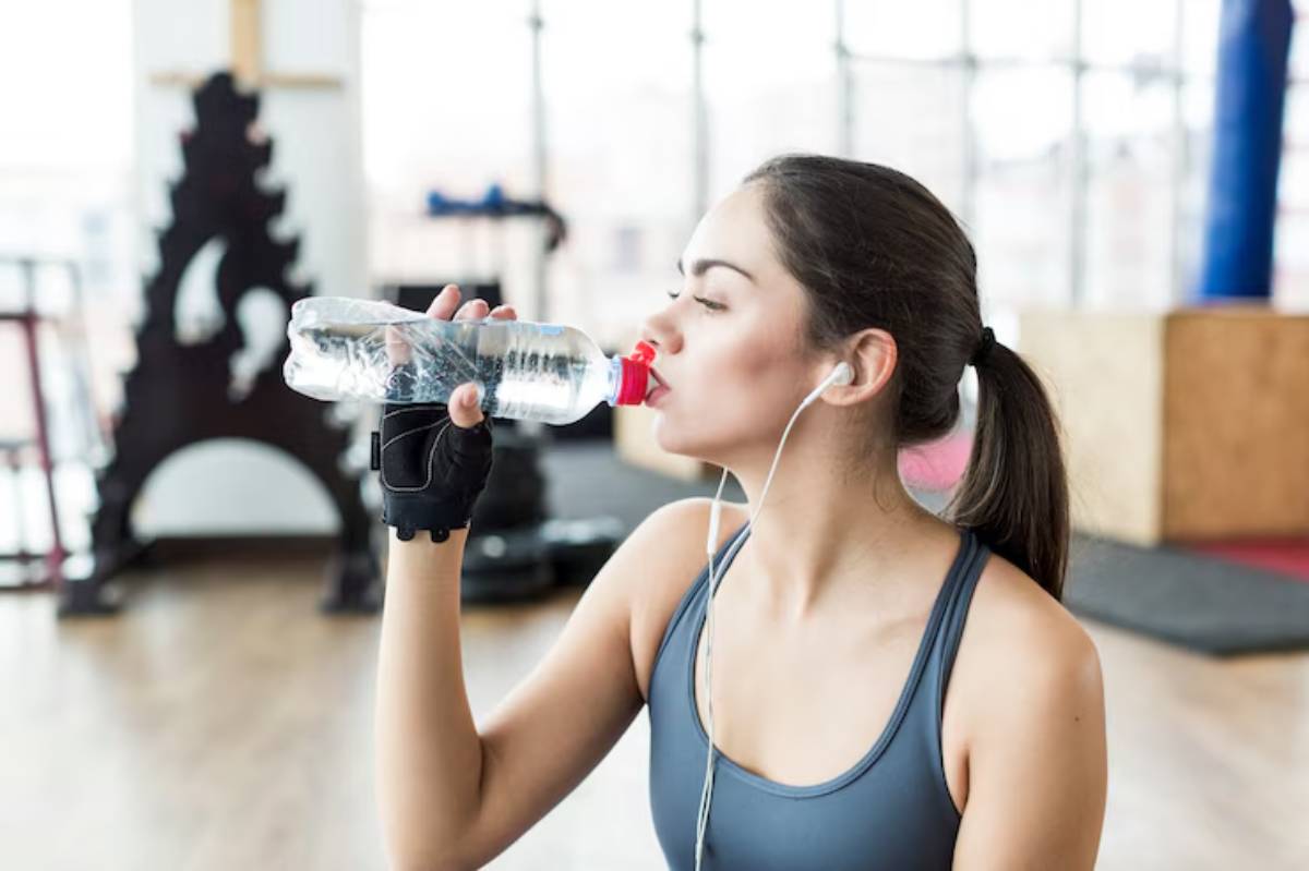 A woman in athletic wear drinks from a plastic water bottle while listening to music in a gym setting.