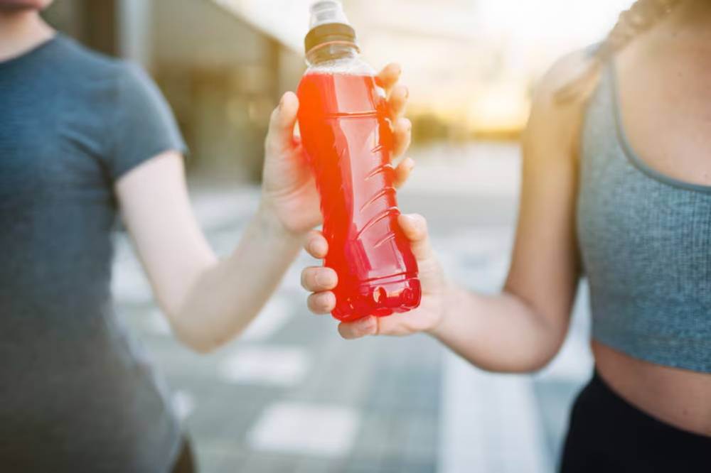 Two individuals hold a bright red sports drink bottle together, set against an outdoor background during golden hour.