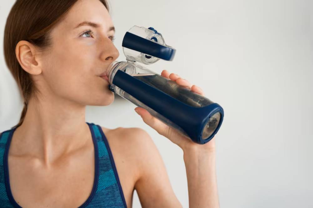 A person holds a blue and transparent fitness water bottle, wearing a sports tank top, with a plain wall background.