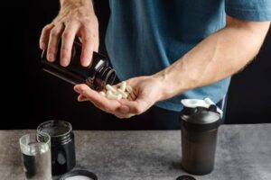 A person pours supplements from a black bottle into their palm, with a shaker and glass of water on the table.