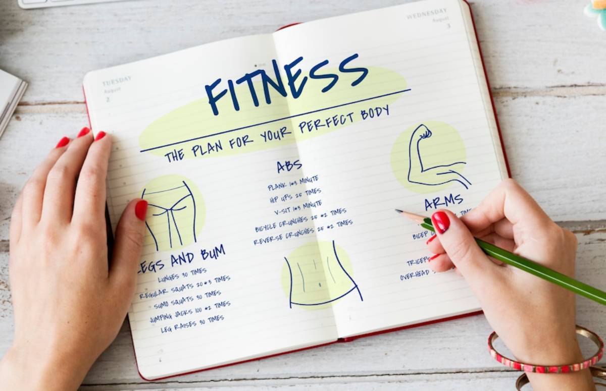 A person's hand notes a fitness plan in a journal, detailing exercises for abs, arms, and legs with illustrations and instructions.