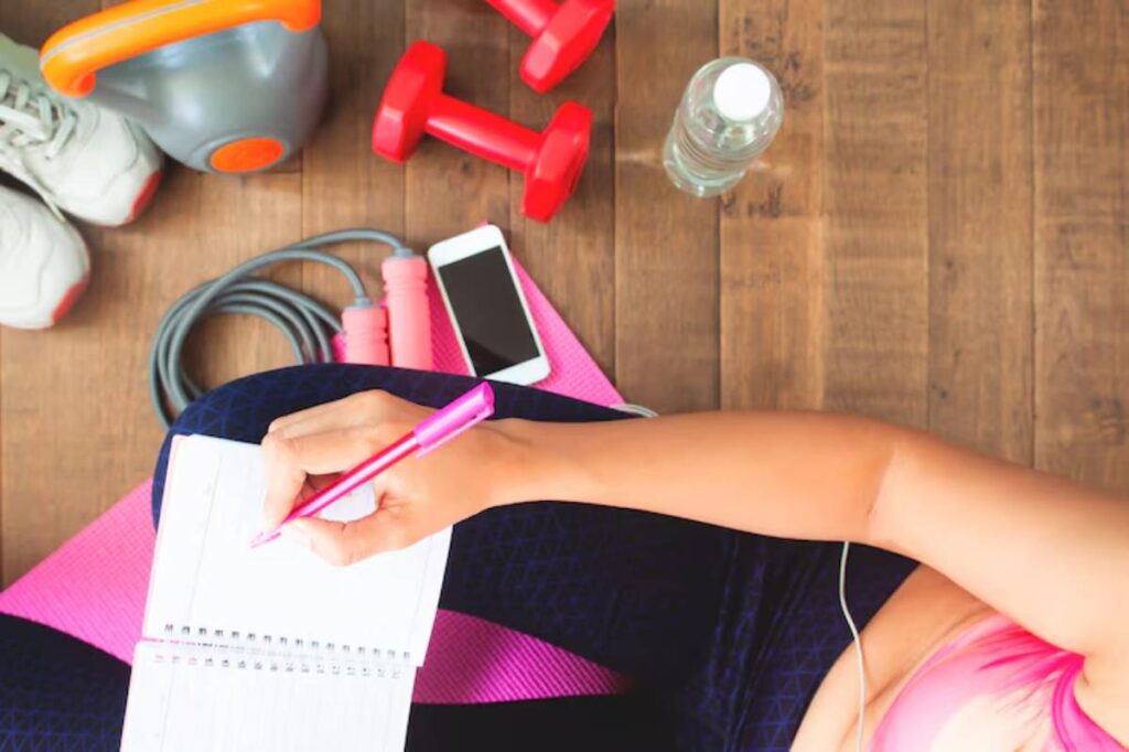 A person writes in a notebook while sitting on a yoga mat surrounded by workout gear, including dumbbells, a kettlebell, and a water bottle.