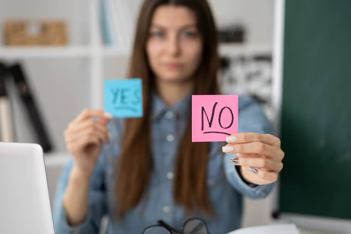 A person holds two sticky notes, one blue with 'YES' and one pink with 'NO,' in front of a computer and a green chalkboard.