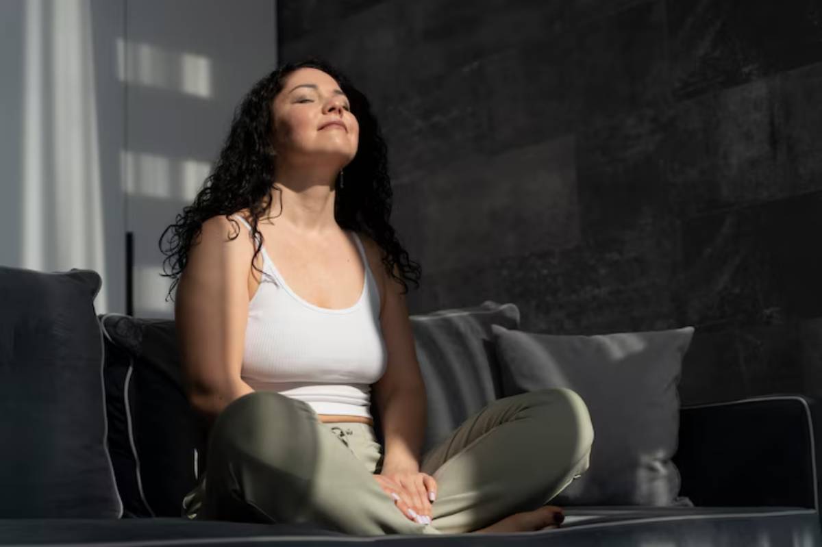 A person with curly hair sits cross-legged on a couch and doing mindfulness, wearing a white tank top and light green pants, bathed in soft, natural light.