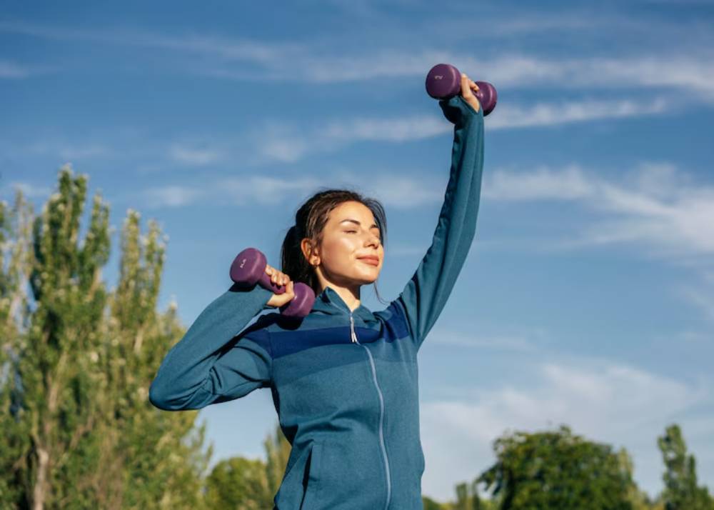A person in a blue jacket is exercising outdoors, lifting purple dumbbells against a clear blue sky and greenery in the background.