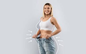 A woman in a white tank top holds oversized jeans, showcasing her slim waist and fit physique against a light gray background.