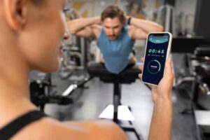 A person holds a smartphone displaying a timer at 45 seconds, while another person performs an exercise on a bench in a gym.