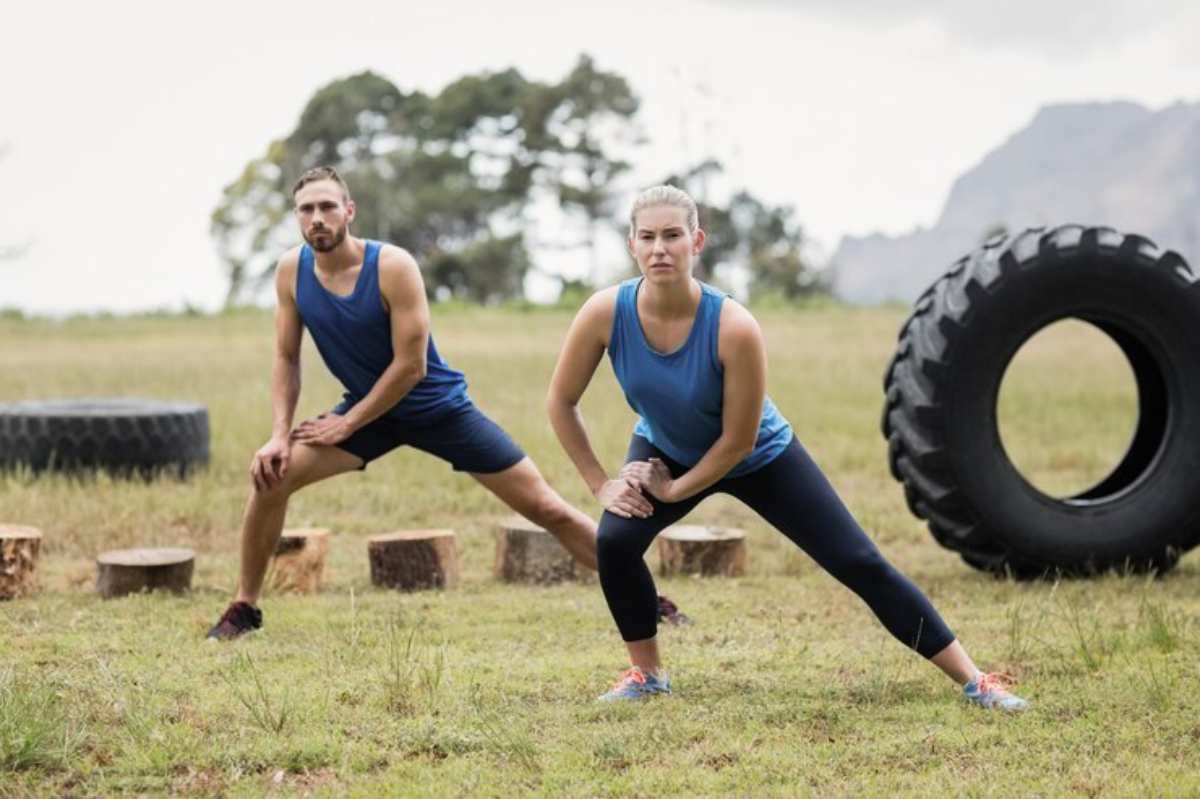 Two individuals in blue athletic tops perform side lunges outdoors, surrounded by grass, wooden blocks, and a large tire.