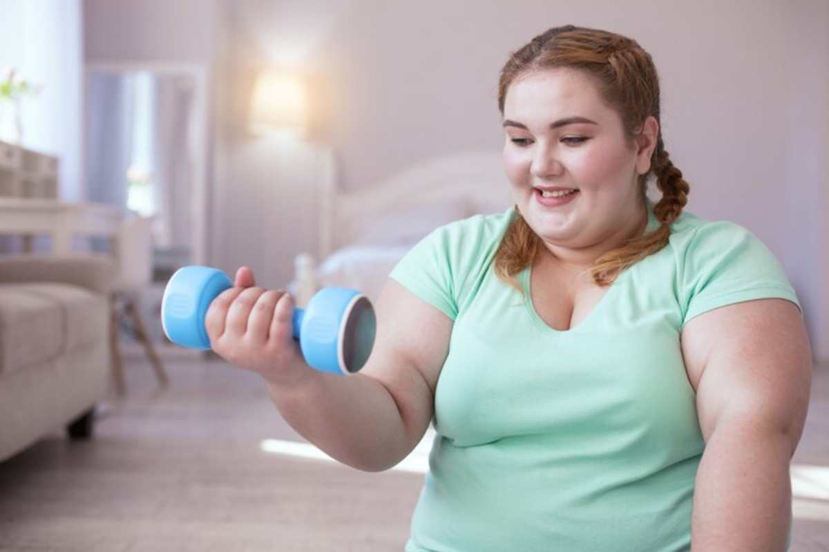 A person in a light green shirt holds a blue dumbbell, exercising in a bright, modern living room.
