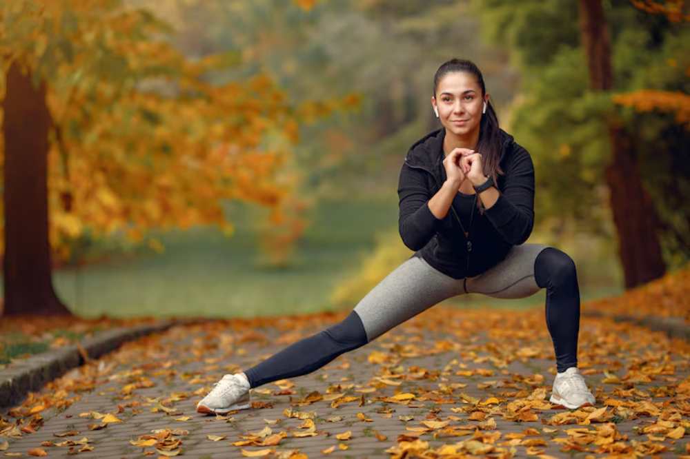 A person in athletic wear performs a stretching exercise on a cobblestone path surrounded by autumn foliage.