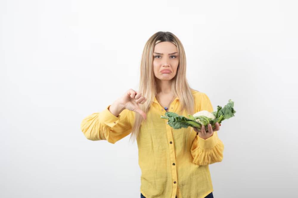 A woman in a yellow shirt holds a piece of cauliflower and gives a thumbs down gesture, expressing dislike for the vegetable.